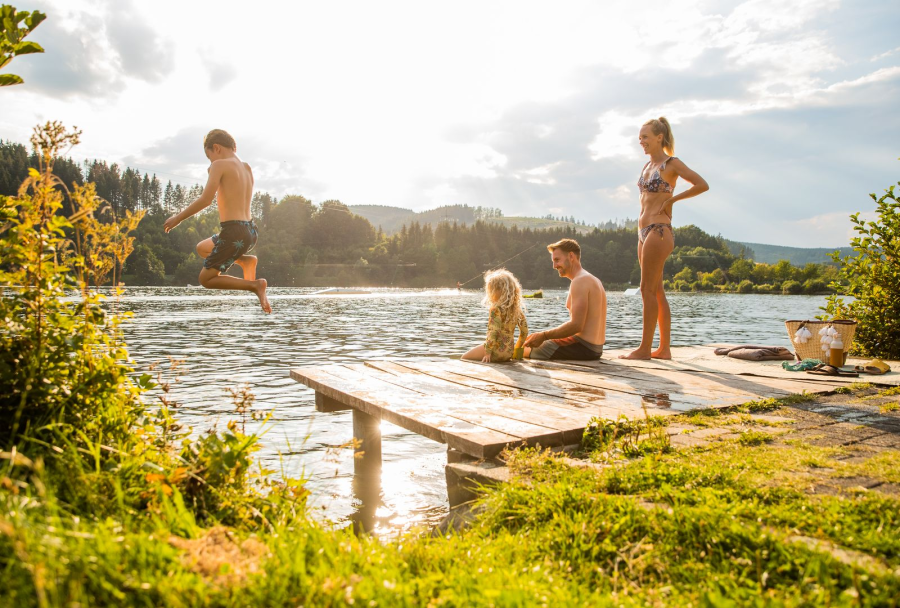 Eine Familie sitzt auf einem Steg am Ufers, während eines der Kinder ins Wasser springt